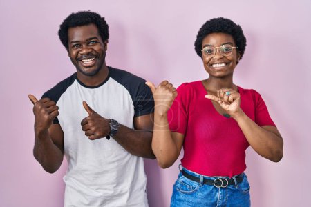 Foto de Joven pareja afroamericana de pie sobre fondo rosa apuntando a la espalda con la mano y los pulgares hacia arriba, sonriendo confiado - Imagen libre de derechos