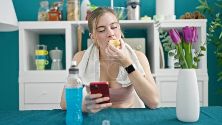 Foto de Mujer rubia joven usando ropa deportiva comiendo manzana usando teléfono inteligente en el comedor - Imagen libre de derechos
