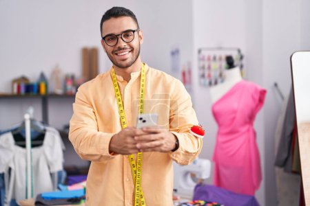 Téléchargez les photos : Jeune homme hispanique tailleur souriant confiant en utilisant smartphone au magasin de tailleur - en image libre de droit