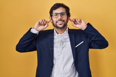 Foto de Hombre latino guapo de pie sobre fondo amarillo sonriendo tirando de las orejas con los dedos, gesto divertido. problema de audición - Imagen libre de derechos