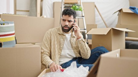 Photo for Young hispanic man talking on smartphone unpacking cardboard box at new home - Royalty Free Image