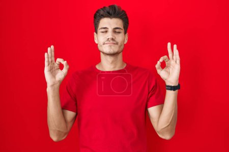 Téléchargez les photos : Jeune homme hispanique debout sur fond rouge détendu et souriant les yeux fermés faisant geste de méditation avec les doigts. concept de yoga. - en image libre de droit