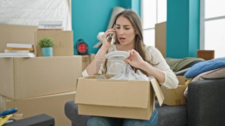 Photo for Young beautiful hispanic woman unpacking cardboard box speaking on the phone complaining at new home - Royalty Free Image