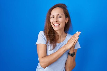 Téléchargez les photos : Brunette femme debout sur fond bleu applaudissements et applaudissements heureux et joyeux, souriant mains fières ensemble - en image libre de droit