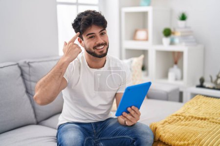 Foto de Hombre hispano con barba usando touchpad sentado en el sofá sonriendo apuntando a la cabeza con un dedo, gran idea o pensamiento, buena memoria - Imagen libre de derechos