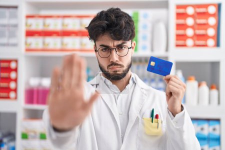 Téléchargez les photos : Homme hispanique avec barbe travaillant à la pharmacie pharmacie tenant la carte de crédit avec la main ouverte faisant stop signe avec expression sérieuse et confiante, geste de défense - en image libre de droit