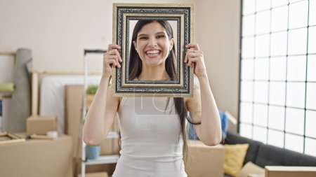 Foto de Joven hermosa mujer hispana sonriendo confiada sosteniendo marco en nuevo hogar - Imagen libre de derechos