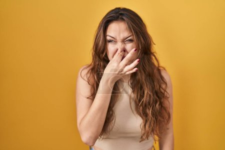 Photo for Young hispanic woman standing over yellow background smelling something stinky and disgusting, intolerable smell, holding breath with fingers on nose. bad smell - Royalty Free Image
