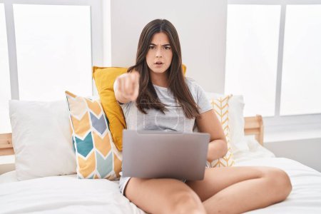 Téléchargez les photos : Femme hispanique utilisant ordinateur portable sur le lit pointant du doigt à la caméra et à vous, geste confiant regardant sérieux - en image libre de droit