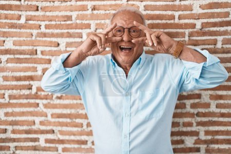 Photo for Senior man with grey hair standing over bricks wall doing peace symbol with fingers over face, smiling cheerful showing victory - Royalty Free Image