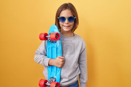 Foto de Pequeño niño hispano con gafas de sol sosteniendo el patín con un aspecto positivo y feliz de pie y sonriendo con una sonrisa confiada mostrando los dientes - Imagen libre de derechos