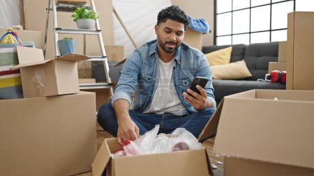 Photo for African american man unpacking cardboard box checking with smartphone at new home - Royalty Free Image