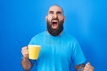 Téléchargez les photos : Jeune homme hispanique avec barbe et tatouages boire une tasse de café en colère et fou hurlant frustré et furieux, criant de colère. rage et concept agressif. - en image libre de droit