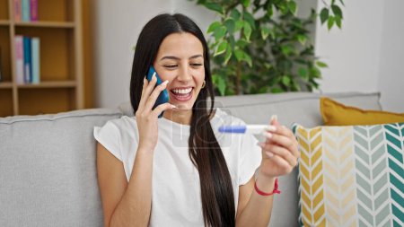 Photo for Young beautiful hispanic woman talking on smartphone holding pregnancy test at home - Royalty Free Image