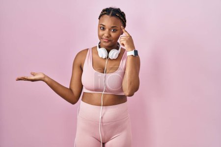 Photo for African american woman with braids wearing sportswear and headphones confused and annoyed with open palm showing copy space and pointing finger to forehead. think about it. - Royalty Free Image
