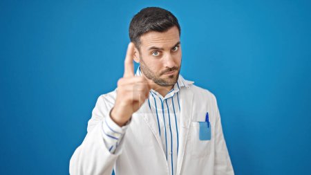 Foto de Joven médico hispano de pie con expresión infeliz señalando con el dedo a la cámara sobre un fondo azul aislado - Imagen libre de derechos