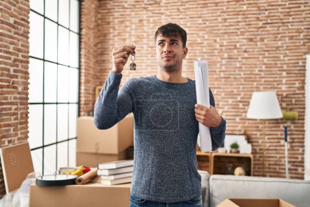 Photo for Young hispanic man holding keys of new home and blueprints smiling looking to the side and staring away thinking. - Royalty Free Image