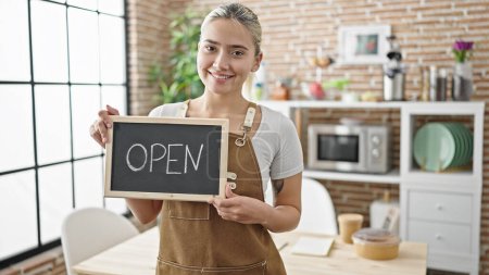 Photo for Young beautiful hispanic woman wearing apron holding open blackboard at dinning room - Royalty Free Image