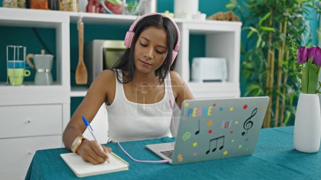 Téléchargez les photos : Femme afro-américaine en utilisant ordinateur portable et écouteurs écriture sur ordinateur portable à la salle à manger - en image libre de droit