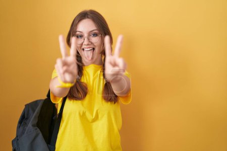 Téléchargez les photos : Jeune femme caucasienne portant un sac à dos étudiant sur fond jaune souriant avec la langue montrant les doigts des deux mains faisant signe de victoire. numéro deux. - en image libre de droit