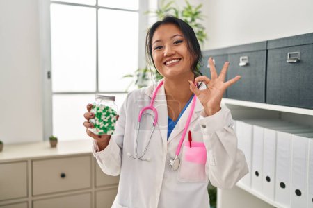 Foto de Joven asiática médico mujer sosteniendo píldoras haciendo ok signo con los dedos, sonriendo amable gesto excelente símbolo - Imagen libre de derechos