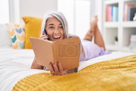 Photo for Middle age grey-haired woman talking on smartphone reading book at bedroom - Royalty Free Image
