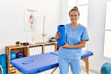 Photo for Young beautiful hispanic woman physiotherapist smiling confident holding foam roller at rehab clinic - Royalty Free Image
