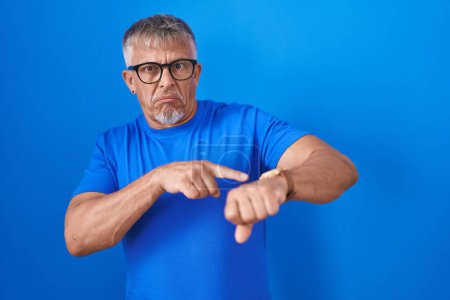 Téléchargez les photos : Homme hispanique avec les cheveux gris debout sur fond bleu à la hâte pointant vers regarder le temps, l'impatience, bouleversé et en colère pour retard de délai - en image libre de droit