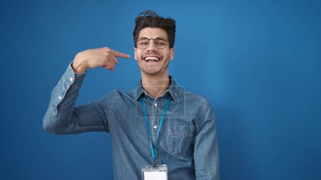 Téléchargez les photos : Jeune homme hispanique souriant confiant pointant du doigt vers la bouche sur fond bleu isolé - en image libre de droit