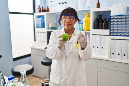 Foto de Mujer hispana trabajando en laboratorio científico con manzana sonriendo mirando al costado y mirando hacia otro lado pensando. - Imagen libre de derechos