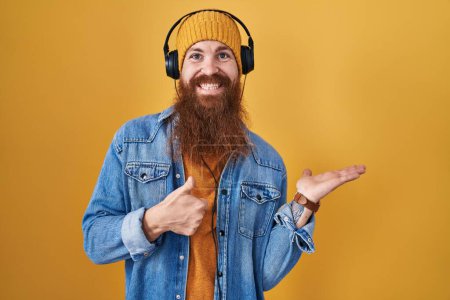 Foto de Hombre caucásico con barba larga escuchando música usando auriculares mostrando la palma de la mano y haciendo buen gesto con los pulgares hacia arriba, sonriendo feliz y alegre - Imagen libre de derechos