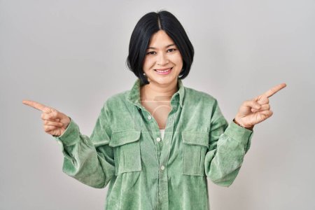 Foto de Joven mujer asiática de pie sobre fondo blanco sonriendo confiado señalando con los dedos a diferentes direcciones. espacio de copia para publicidad - Imagen libre de derechos