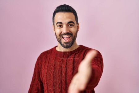 Foto de Joven hombre hispano con barba vistiendo suéter casual sobre fondo rosa sonriente amistoso ofreciendo apretón de manos como saludo y bienvenida. negocios exitosos. - Imagen libre de derechos