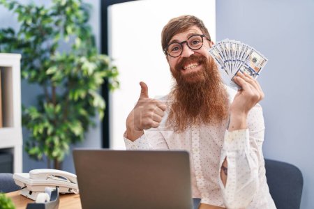 Foto de Hombre caucásico con barba larga trabajando con computadora portátil sosteniendo dólares sonriendo feliz y positivo, pulgar hacia arriba haciendo excelente y signo de aprobación - Imagen libre de derechos