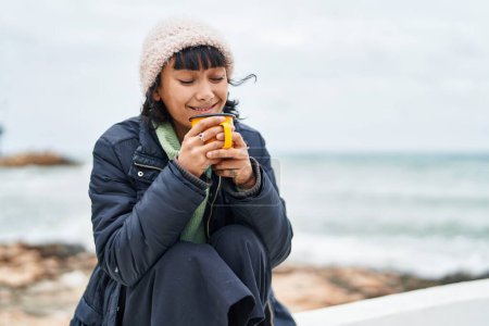 Foto de Joven hermosa mujer hispana sonriendo confiada bebiendo café en la playa - Imagen libre de derechos