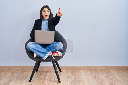 Téléchargez les photos : Jeune femme hispanique assise sur une chaise à l'aide d'un ordinateur portable pointant du doigt surpris devant, bouche ouverte expression étonnée, quelque chose sur le devant - en image libre de droit