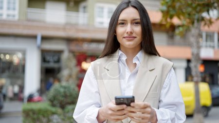Photo for Young beautiful hispanic woman using smartphone smiling at street - Royalty Free Image