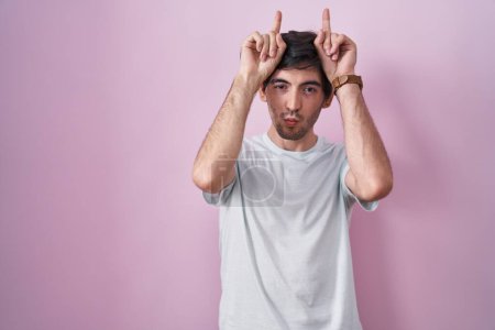 Téléchargez les photos : Jeune homme hispanique debout sur fond rose faisant un geste drôle avec le doigt sur la tête comme des cornes de taureau - en image libre de droit