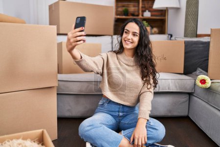 Photo for Young hispanic woman make selfie by the smartphone sitting on floor at new home - Royalty Free Image