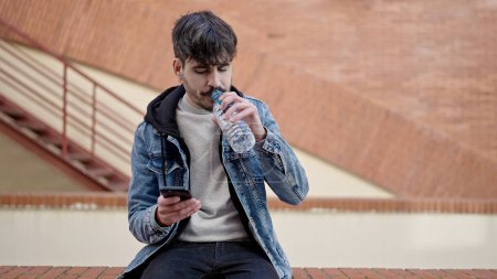 Téléchargez les photos : Jeune homme hispanique assis eau potable à l'aide d'un smartphone dans la rue - en image libre de droit