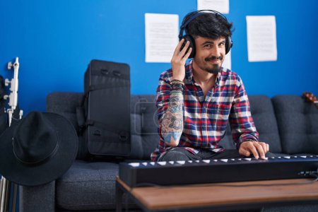 Foto de Joven músico hispano sonriendo confiado tocando el teclado del piano en el estudio de música - Imagen libre de derechos