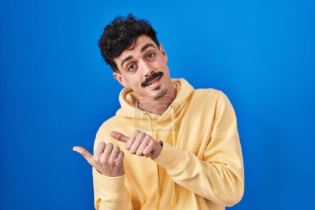 Foto de Hispanic man standing over blue background pointing to the back behind with hand and thumbs up, smiling confident - Imagen libre de derechos