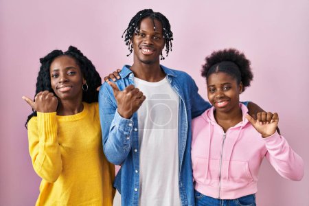 Foto de Grupo de tres jóvenes negros de pie juntos sobre fondo rosa apuntando hacia atrás con la mano y los pulgares hacia arriba, sonriendo confiados - Imagen libre de derechos