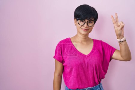 Foto de Joven mujer asiática con el pelo corto de pie sobre fondo rosa sonriendo mirando a la cámara mostrando los dedos haciendo signo de victoria. número dos. - Imagen libre de derechos