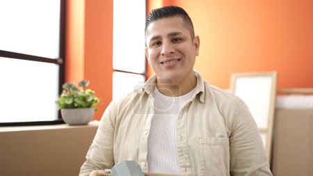 Foto de Joven hispano sonriendo confiado sosteniendo paquete en nuevo hogar - Imagen libre de derechos