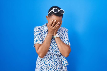 Foto de Mujer afroamericana de pie sobre fondo azul con expresión triste cubriendo la cara con las manos mientras llora. concepto de depresión. - Imagen libre de derechos