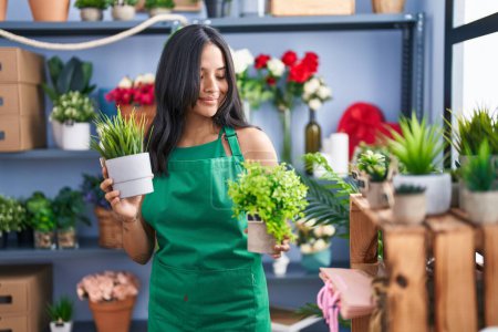 Téléchargez les photos : Jeune femme hispanique fleuriste souriant confiant tenant des plantes à la boutique de fleuriste - en image libre de droit