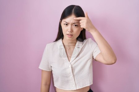 Foto de Mujer joven china de pie sobre fondo rosa señalando infeliz a grano en la frente, fea infección de la espinilla. acné y problemas de piel - Imagen libre de derechos