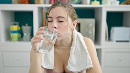 Photo for Young blonde woman wearing sportswear drinking glass of water at dinning room - Royalty Free Image