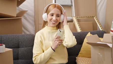 Foto de Young blonde woman listening to music sitting on sofa at new home - Imagen libre de derechos